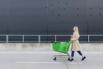 woman with blond hair in a raincoat rolls a shopping cart against a wall