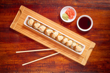 Wooden Table Served with Asian Food Consisting of Sushi with Crab Stuffing and Tempura around Rice with Paprika Mayonnaise Accompanied by Soya Sauce, Pickled Ginger, Wasabi and Sticks
