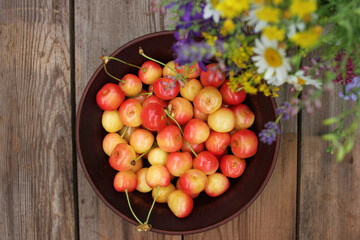 Naklejka na ściany i meble Yellow and pink cherries in a bowl, and midsummer wild flowers on a vintage wooden board background. Summer composition