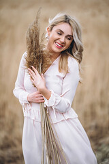 Photo of a pretty smiling girl with long blond curly hair in light long drees standing in a reed field