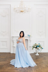 Young pregnant woman in a light classic interior. Caucasian woman with black hairs in long romantic dress near the blue sofa. Concept of new life. Waiting of baby. Pretty pregnant woman in a room