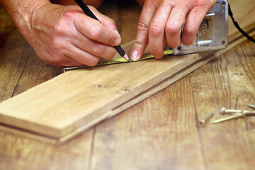 close-up of hands making a mark with a pencil on a wooden board on a wooden workbench in a craftsman's workshop, screws on the floor, illustrating the concept made by yourself and the DIY courses
