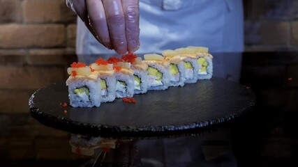 Chef is decorating sushi maki rice rolls with flying fish roe, tobiko caviar. Sushi maki cntains salmon, mango, cream cheese, lemon. Appetizing fusion roll. Restaurant menu. 