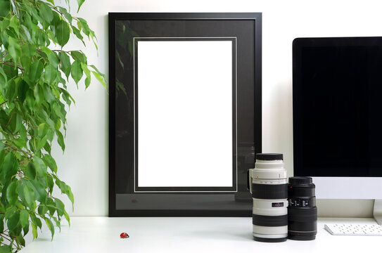 Empty frame to personalize placed on a white desk next to a computer, camera lens and a potted ficus tree, for staging a photo or drawing