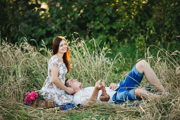 Happy couple of boyfriend and girlfriend sitting among grass in meadow, at picnic. Man lying on knees of his wife and looking up. Woman in dress in floral print smiling and dreaming. Summertime.