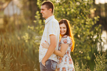 Side view of couple of handsome man and brunette woman with long hair posing and standing among grass in meadow. Girlfriend embracing her boyfriend from back and looking at camera, Sunny summer day.