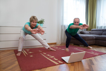 active senior couple doing stretching exercise and watching online workout tutorials on the laptop in living room at home.  home fitness, activewear.