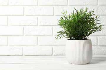 Beautiful artificial plant in flower pot on white wooden table near brick wall. Space for text
