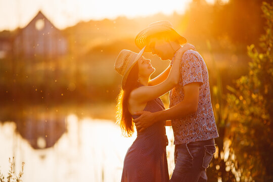 Portrait of couple of brunette woman and man standing on pier of lake at sunset, embracing and smiling. Girlfriend and boyfriend sensuality looking each other. Girl in dress, man in shirt and hat.