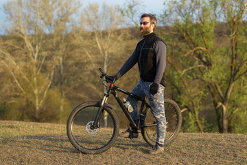 Sports brutal bearded guy on a modern mountain bike. Cyclist on the green hills in the spring.