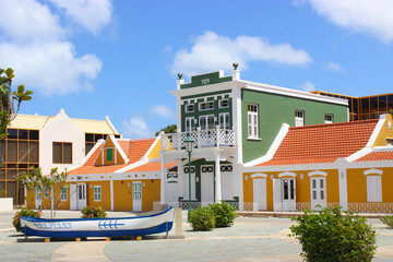 Several colored buildings stand on the street. The sun shines brightly, the sky is blue. In the foreground on the ground is a white-blue boat. Aruba August 2014 - obrazy, fototapety, plakaty