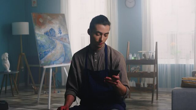 Portrait of young artist man sitting on a stool in apron and using his mobile phone, gadget, creative bright studio, canvas, tools, and elements of decor are on the background, Zoom In, Slow motion.