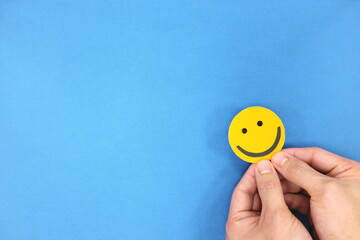 Happiness and positivity concept. Hand holding yellow smiling face in blue background with copy...