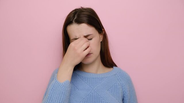 Sad tired of glasses young woman massaging nose bridge, feeling eye strain, exhausted lady suffering from headache or migraine, wears blue sweater, isolated on pink background. Eyes fatigue concept