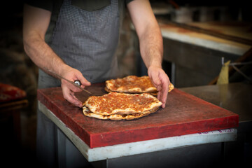 Baked lahmacun is a round, thin piece of dough topped with minced meat (most commonly beef or lamb), minced vegetables and herbs. Man cuts lahmacun.