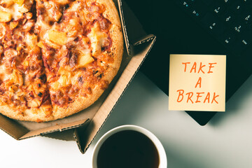 Pizza in box, cup of coffee, laptop and sticker with text TAKE A BREAK on table in office.