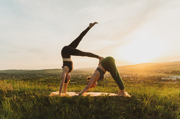 Two young caucasian women practicing partner yoga in on a hill at sunset down dog and one legged-handstand