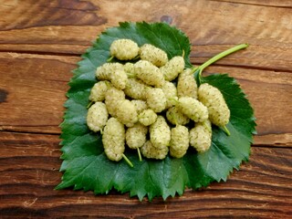 White mulberry fruit on green mulberry leaf. Fresh ripe white mulberry berries & leaf on wooden table background. Organic white berries (Morus alba) from tree garden for juice or jam - harvest concept