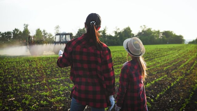 Family farmers work in the field, look at the digital tablet against the background of the tractor. The farmer mother and her daughter talk in the field, use a tablet. Smart and modern farm concept.