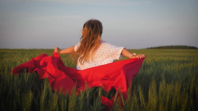 Young girl running with red tissue in green field. Happy cute girl playing in the wheat field on a warm summer day at sunset.