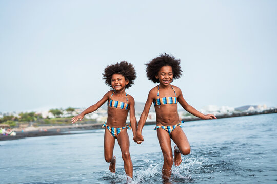 Two sisters running inside the water on the beach during summer time - Happy kids having fun on vacation time - Travel, love and family concept - Focus on faces