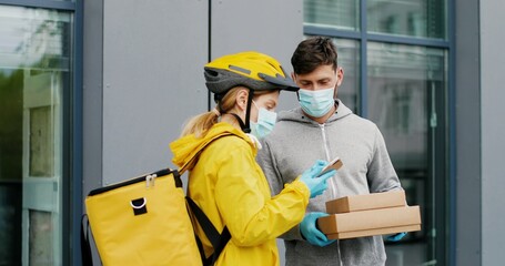 Caucasian young female courier in casque, gloves and medical mask giving carton parcels to man at street. Woman byciclist with bag bringing boxes to male customer outdoors.