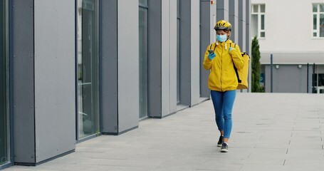 Portrait of female courier in medical mask and helmet for riding bike with yellow thermal bag for food delivery using smartphone. Food delivery concept