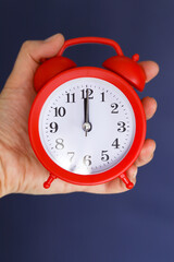 Red beautiful alarm clock with black arrows in the hand of a Caucasian man. Selective focus. The hand on the clock shows midnight or noon.