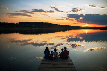Naklejka na ściany i meble family of four people sitting on the shore on a wooden bridge of a large lake in summer and watching the beautiful sunset back view