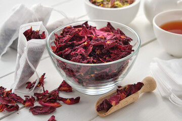 Bowl of dry rose petals, tea bags filled with red petals of heathy rose. Herbal tea cup, teapot and mortar on wooden table.