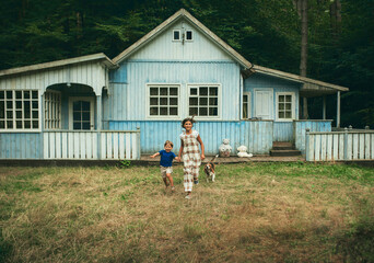 Fototapeta na wymiar Children, a boy and a girl with a dog joyfully run near their wooden blue house in Scandinavian style, in summer, along the grass