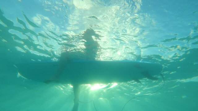 SLOW MOTION UNDERWATER: Unrecognizable female surfer sits on her longboard in line up and waits for big waves on a sunny summer day. Woman on vacation in Barbados sits in line up, waiting for waves.