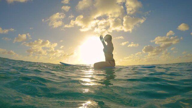 SLOW MOTION, LOW ANGLE, LENS FLARE, CLOSE UP Young Caucasian woman on awesome surfing trip in Barbados sits on her surfboard at sunset and splashes the refreshing water while waiting in line up.