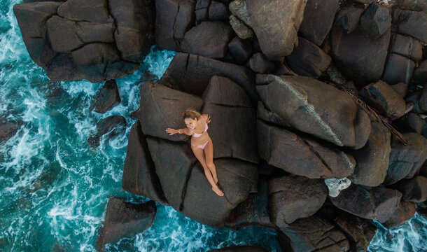 Beautiful slim blonde young woman in nude bikini sitting on rocks. Drone aerial shot. Picturesque ocean landscape with girl from above. Summer holiday in Sri Lanka