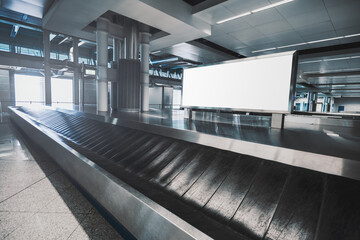 A white blank rectangular advertising billboard mock-up in airport baggage claim hall with a conveyor in front; advert poster template indoors; a white empty banner mockup near a luggage transporter