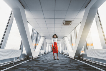 Wide-angle view of a stately African-American businesswoman in a red skirt and with a cellphone in hand standing in the middle of a passageway between two towers of a modern business skyscrapers