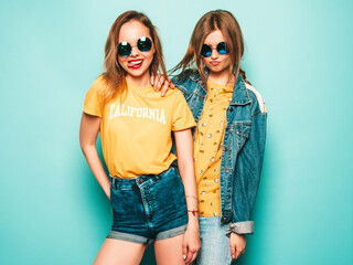 Two young beautiful smiling hipster girls in trendy summer yellow T-shirts and jeans jacket. Sexy carefree women posing near blue wall. Trendy and positive models having fun