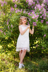 Young woman in blooming near lilac bush in a garden. Spring story. Romantic look with white lace dress. Happy girl in beautiful spring day. Pretty slim blonde woman, caucasian