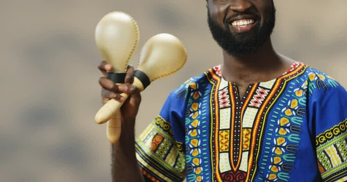Handsome young African American man in traditional clothes smiling cheerfully and playing on maracas. Close up of happy guy holding musical instruments and making rhytm to music while shaking.