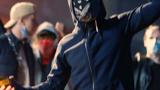 Caucasian young male protester holding Molotov cocktail and jumping in aggressive crowd at protest against police violence. Man in mask on face with fire bottle shouting in rebellion.