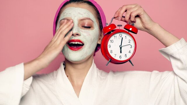 Sleeping girl with mask on face holds alarm clock in hand. Beautiful girl with cosmetic mask on face. Woman wearing face mask. Face care concept.