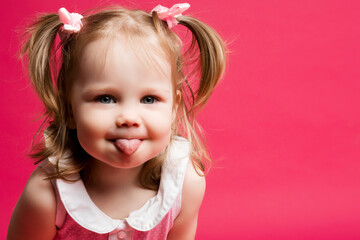 Portrait of funny cute little blonde girl with two tails showing tongue and making face at camera. Sweet child with blue eyes, in pink dress playing at studio. Pink studio background.