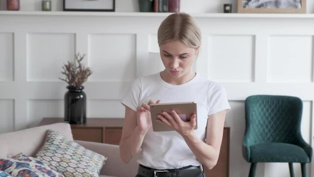 Woman scrolling tablet screen and smiling while standing in the modern room. 