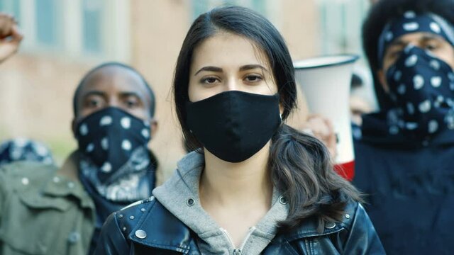 Close up of young Caucasian beautiful woman in mask looking straight at camera outdoor at street riot. Portrait of girl with male Afrcan Americans on background. Female protester at manifestation.