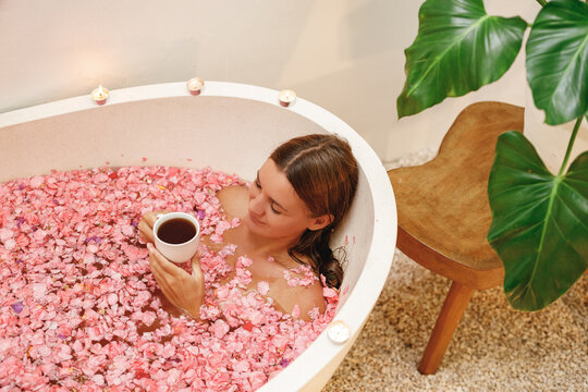 Young Happy Woman Lie In Bath Tub Full Of Pink Roses Flower Petals Drink Herbal Tea, Enjoy Her Day In The Spa. White Bath Tube With Pink  Tropical Flowers Petals, Spa, Relaxation, Body Care, Therapy