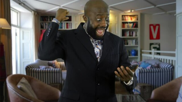 Happy Attractive Young Male Is Talking On The Phone At Home. The Man Receives Good News. The Bearded African Man  Is Smiling, Chatting And Celebrating In His Living Room.