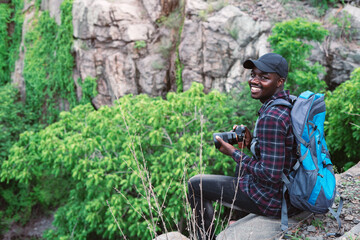 African man traveler sitting on the cliff and holding camera with backpack