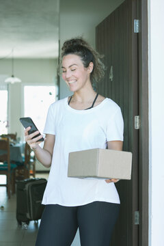 Beautiful Caucasian Young Woman Smiling Receiving Mail Delivery Package At Home During Covid 19 Pandemic. Smartphone Track App