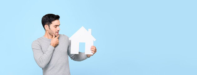 Young man looking at house model and thinking isolated on light blue banner background with copy...