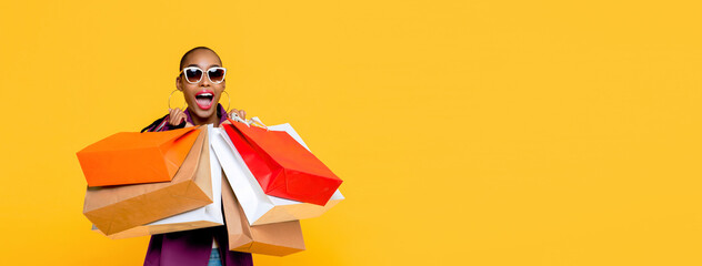Beautiful fashionable African American woman with colorful shopping bags in surprised sale concept isolated on yellow banner background